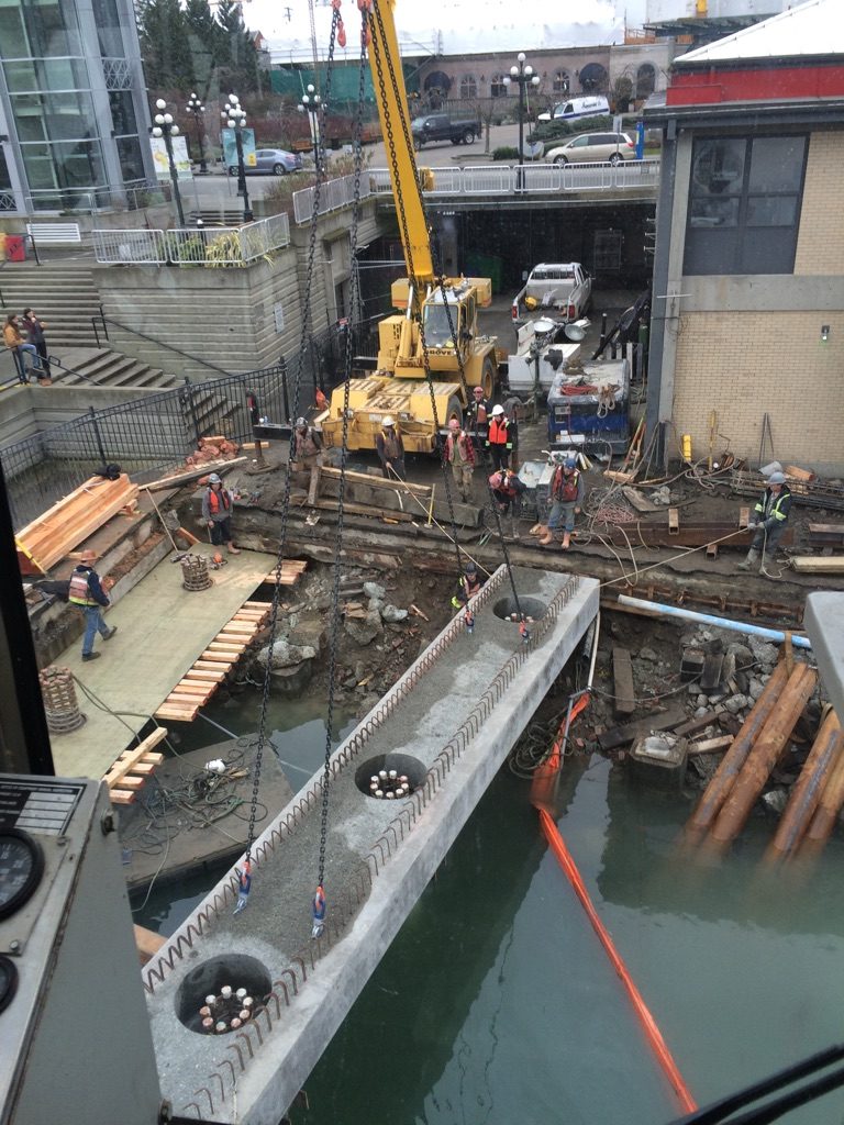 The site of the project is seen with a slab of pre-cast concrete in the middle. There is a crane seen along with some workers on the site. The water can be seen on the lower level of the project site.