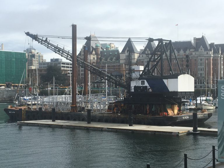 A further view of the construction site with a crane in the middle. It seems to be loading in some steel beams to form the bridge. There is water in the foreground and buildings on the background.
