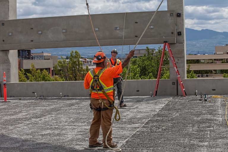 Two men in bright orange workwear can e seen in the middle of the photo which seems to be a floor of the parkade during its construction. They are both in deep focus as one of them pulls over a rope guiding the next precast structure in place.