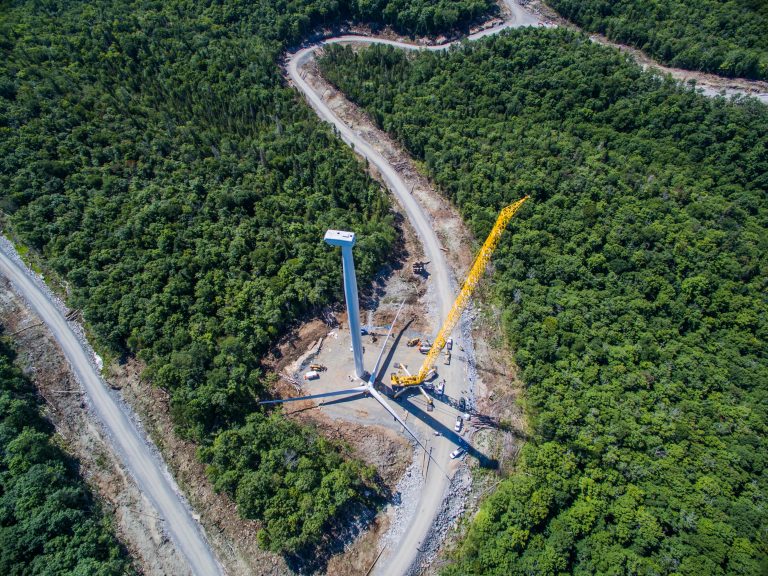 A more aerial view during daytime of the turbine installation. The tower seems to be fully installed now with just the turbine left. There are lots of trees surrounding the wind turbine site.