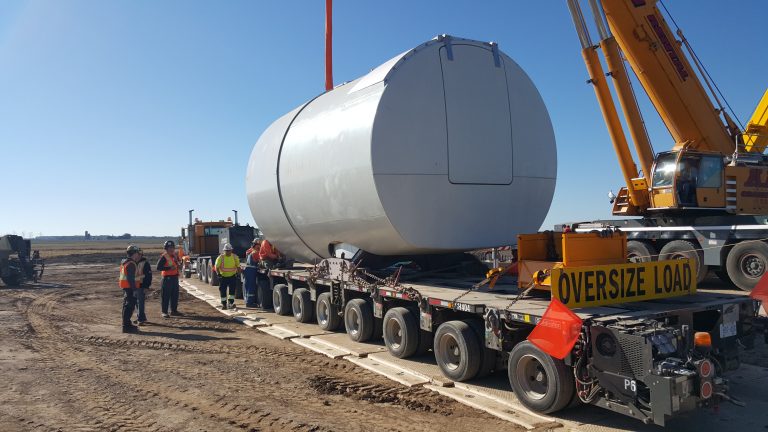 A view of the turbine mechanism being carried by attachments. Some workers can be seen engaged in conversation about the project. It is a clear day.