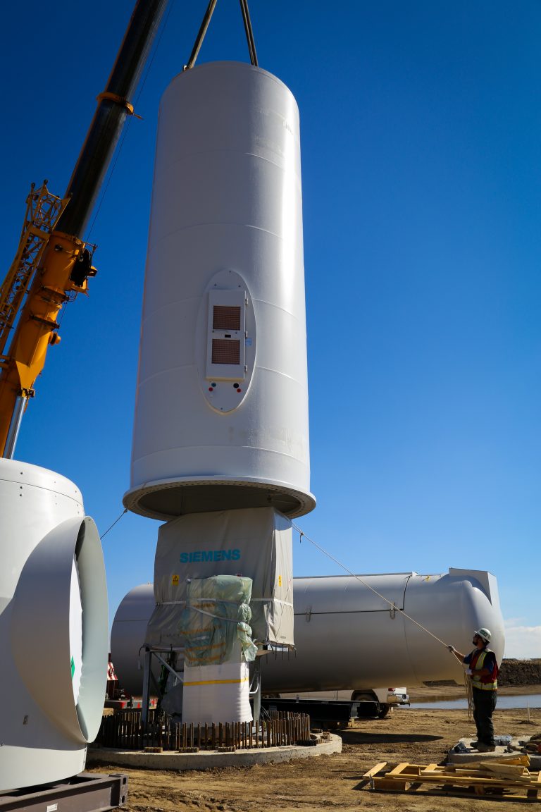 Pieces of the wind turbine body are being hoisted up by the crane as a worker uses a rope to make sure the pieces are aligned.