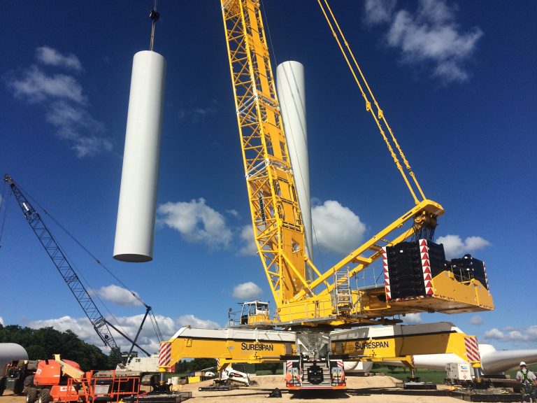A closer side view of the turbine tower being installed with the LG1750 crane. Other trucks can be seen on the site with with a bright blue sky background with some clouds.