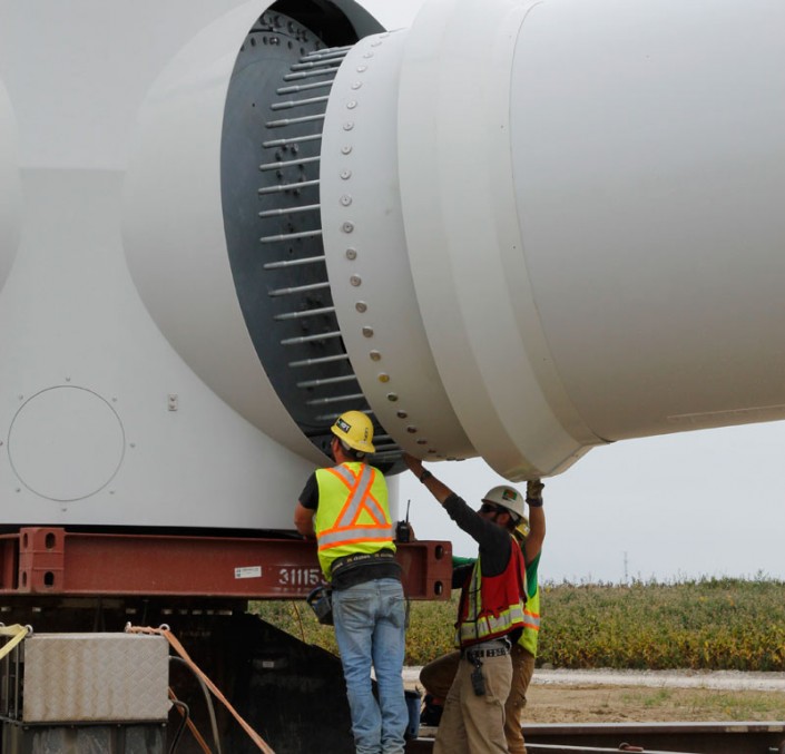 A side view of the turbine as two workers in hard hats do final inspections. There is some grass in the background.