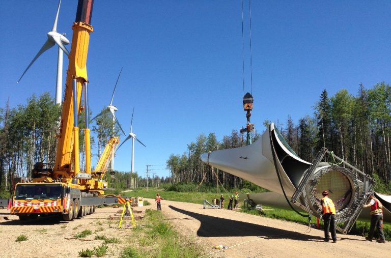 A ground view of the project in motion. A crane can be seen transporting a turbine blade along with some workers guiding the part along. There are tall trees in the background in green on a bright sunny cloudless day.