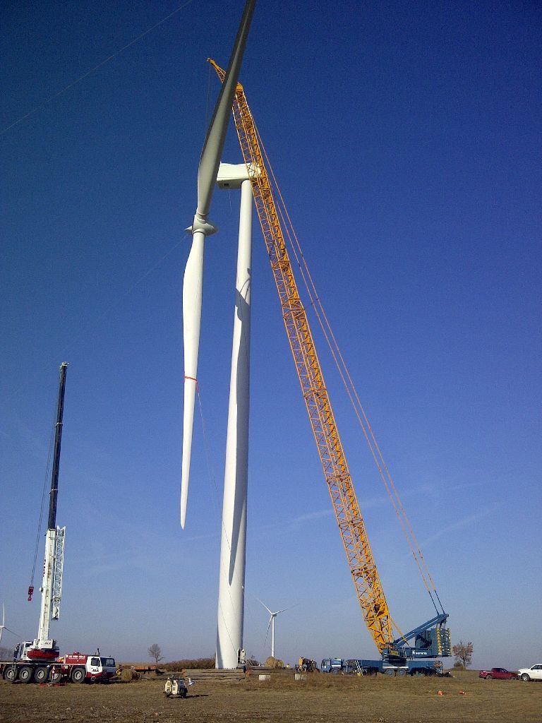 A yellow crane is seen putting in the final touches of the wind turbine after its full erection. Seen on the right is another crane providing support. It is a bright cloudless blue day.